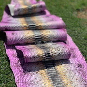 May include: Rolled pink and yellow reptile skins with a scale pattern are stacked on green grass. The skins have a central stripe of grey and black. The text "Hafiz Reptile" is visible on the bottom skin.