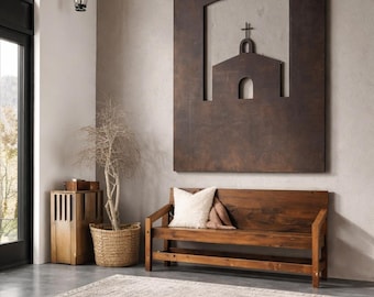 May include: A large rectangular dark brown metal wall art piece featuring a cutout of a church with a cross, positioned above a wooden bench with a white pillow, a woven basket with a plant, and a light-colored area rug with fringe.