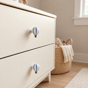 May include: Two drawer knobs shaped like hot air balloons attached to a light cream-coloured dresser. Each knob features a white balloon canopy with vertical blue stripes and a small brown basket at the base, providing a functional handle.