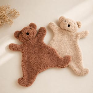 May include: Two flat, plush bear-shaped security blankets made of textured, curly fleece fabric. One bear is dark brown and the other is a light cream colour. Both feature rounded ears, a small snout, and a simple black embroidered nose.