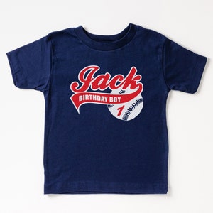 May include: A navy blue short-sleeve t-shirt featuring a graphic of a baseball with red stitching. The text "Jack" is printed in red script above a red banner that reads "BIRTHDAY BOY" in white, with the number "1" in red on the baseball.