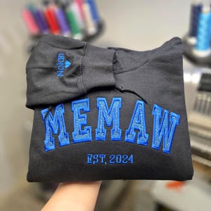 May include: Black hoodie with the word "MEMAW" embroidered in blue, with a glittery outline. The text "EST. 2024" is below the main text. The name "Nolan" is embroidered on the hood in blue.