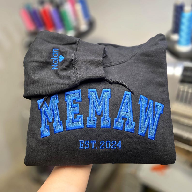 May include: Black hoodie with the word "MEMAW" embroidered in blue, with a glittery outline. The text "EST. 2024" is below the main text. The name "Nolan" is embroidered on the hood in blue.