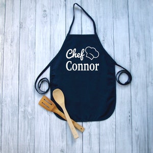 Peut inclure: Un tablier bleu marine avec le texte "Chef Connor" et un dessin de toque de chef blanc. Deux ustensiles de cuisine en bois sont plac&eacute;s sous le tablier. Le tablier a une sangle de cou et des liens &agrave; la taille.