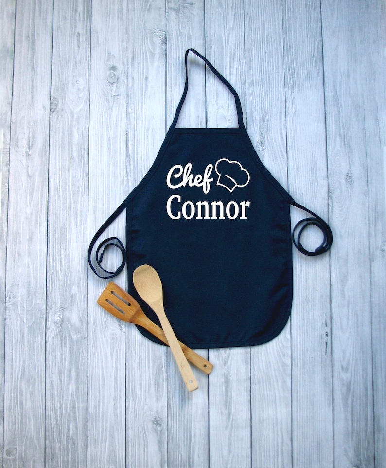 Peut inclure: Un tablier bleu marine avec le texte "Chef Connor" et un dessin de toque de chef blanc. Deux ustensiles de cuisine en bois sont plac&eacute;s sous le tablier. Le tablier a une sangle de cou et des liens &agrave; la taille.