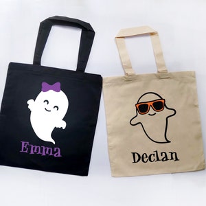 May include: Two Halloween-themed tote bags. One black bag features a white ghost with a purple bow and the name "Emma" in purple. The other is tan with a ghost wearing orange sunglasses and the name "Declan".