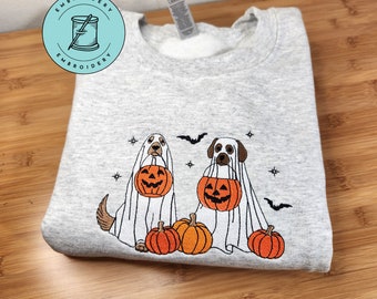 Puede incluir: Una sudadera gris claro de cuello redondo con un bordado de dos perros disfrazados de fantasmas para Halloween. Cada perro sostiene un cubo de calabaza naranja, rodeado de tres calabazas, pequeñas estrellas negras y dos murciélagos.