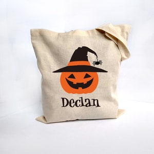 May include: A beige canvas tote bag featuring a Halloween-themed design. The design includes an orange jack-o'-lantern wearing a black witch hat with a spider. The name "Declan" is printed below the pumpkin in black.