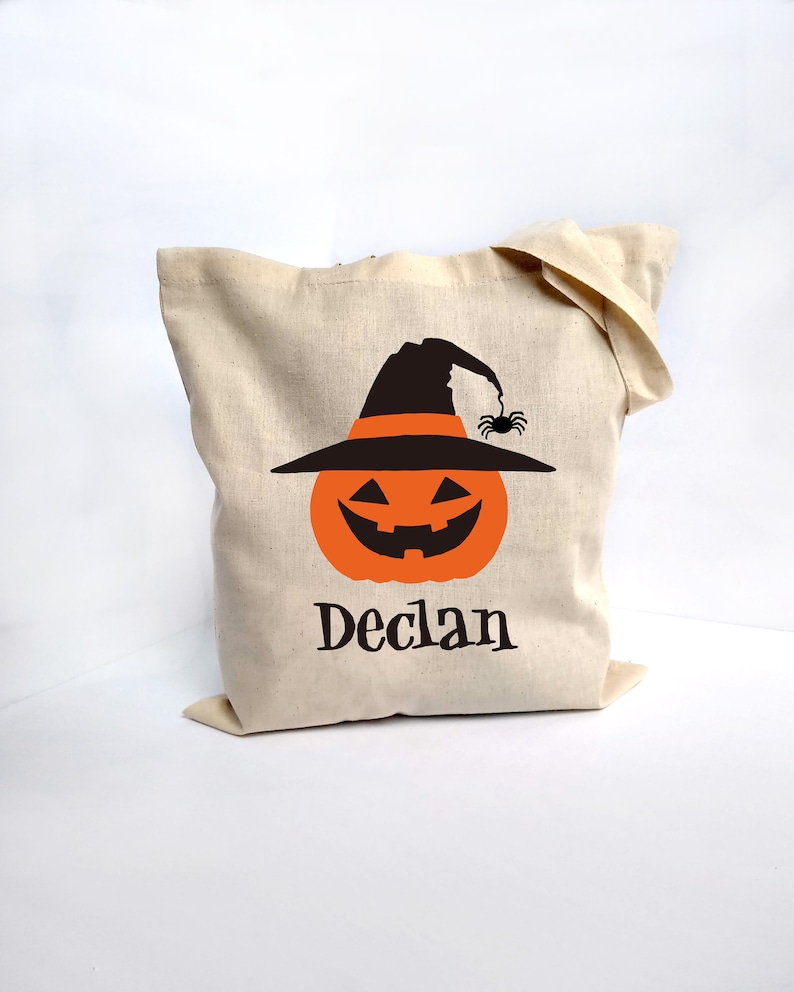 May include: A beige canvas tote bag featuring a Halloween-themed design. The design includes an orange jack-o'-lantern wearing a black witch hat with a spider. The name "Declan" is printed below the pumpkin in black.