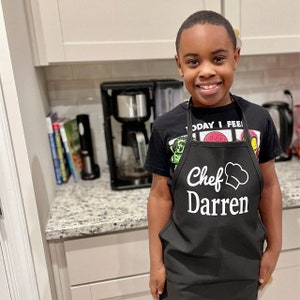 Peut inclure: Un tablier noir avec l'inscription "Chef Darren" en blanc et un dessin de toque de chef. Le tablier est port&eacute; sur un t-shirt noir. L'arri&egrave;re-plan montre une cuisine avec une cafeti&egrave;re.
