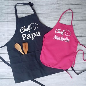 Peut inclure: Deux tabliers de cuisine, un bleu marine et un rose vif, avec du texte blanc et des graphiques de chapeau de chef. Le tablier bleu marine dit "Chef Papa" et a une poche avec des cuill&egrave;res en bois. Le tablier rose dit "Chef Annabelle".