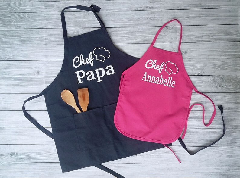 Peut inclure: Deux tabliers de cuisine, un bleu marine et un rose vif, avec du texte blanc et des graphiques de chapeau de chef. Le tablier bleu marine dit "Chef Papa" et a une poche avec des cuill&egrave;res en bois. Le tablier rose dit "Chef Annabelle".