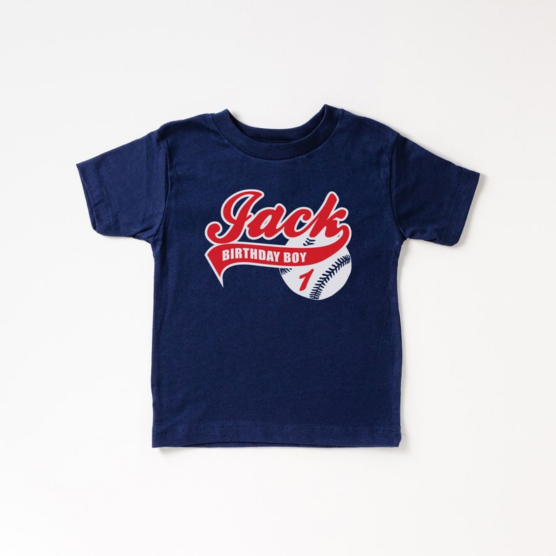 May include: A navy blue short-sleeve t-shirt featuring a graphic of a baseball with red stitching. The text "Jack" is printed in red script above a red banner that reads "BIRTHDAY BOY" in white, with the number "1" in red on the baseball.