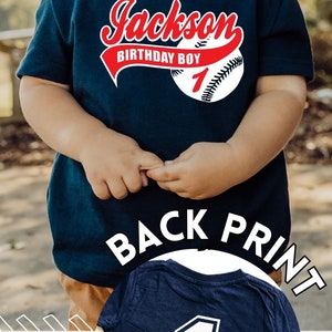 May include: A navy blue short-sleeved t-shirt featuring a graphic of a baseball with the text Jackson, Birthday Boy, and the number 1. An inset shows the back of the shirt, which displays a large white number 1 with a dark blue outline.