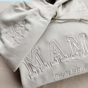 May include: A sand-colored hoodie with ivory thread embroidery. The word "MAM" and "EST. 1996" are embroidered on the front. The left sleeve has embroidered names. The hoodie has a drawstring and a hood.
