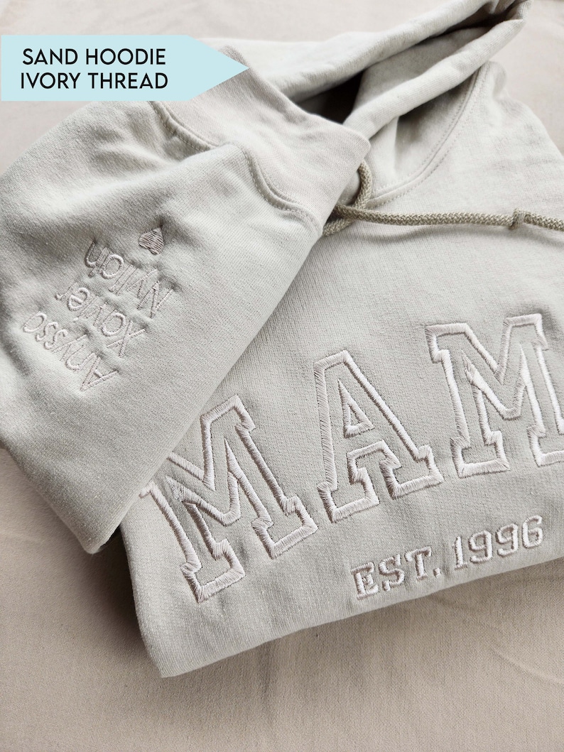 May include: A sand-colored hoodie with ivory thread embroidery. The word "MAM" and "EST. 1996" are embroidered on the front. The left sleeve has embroidered names. The hoodie has a drawstring and a hood.