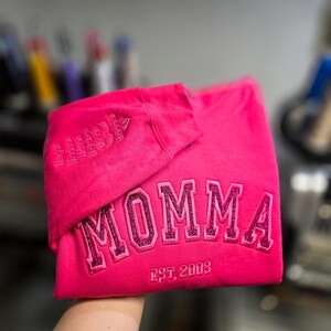 May include: A bright pink sweatshirt with the word "MOMMA" embroidered in glittery pink letters. The text "EST. 2003" is below the word. The sleeve has embroidered names. A casual, comfortable garment.