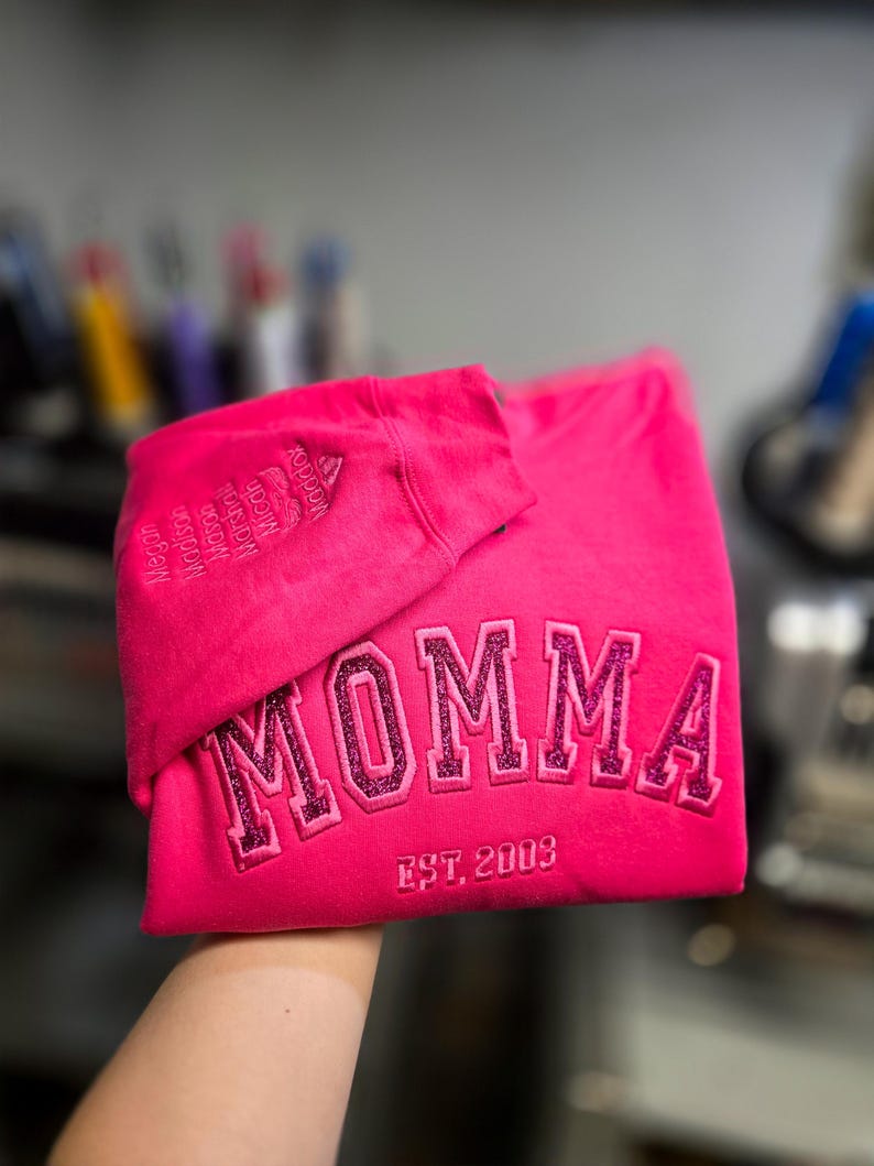 May include: A bright pink sweatshirt with the word "MOMMA" embroidered in glittery pink letters. The text "EST. 2003" is below the word. The sleeve has embroidered names. A casual, comfortable garment.