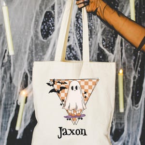 May include: Cream-colored tote bag with a Halloween-themed design. The graphic features a cartoon ghost on a skateboard, set against a triangle with a checkered pattern and bats. The name "Jaxon" is printed below the graphic.