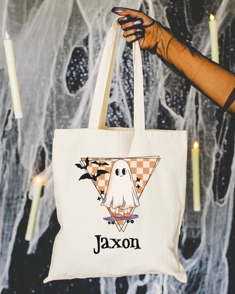 May include: Cream-colored tote bag with a Halloween-themed design. The graphic features a cartoon ghost on a skateboard, set against a triangle with a checkered pattern and bats. The name "Jaxon" is printed below the graphic.