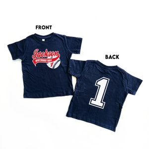 May include: A navy blue short-sleeve t-shirt shown from the front and back. The front features a baseball graphic with the text Jackson BIRTHDAY BOY 1 in red and white. The back displays a large white number 1 with a dark blue outline.