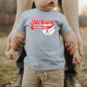 May include: A heather gray short-sleeve t-shirt featuring a graphic of a baseball with red text that reads Jackson, BIRTHDAY BOY, and the number 1. The design is centered on the chest of the shirt, which is worn by a young child.