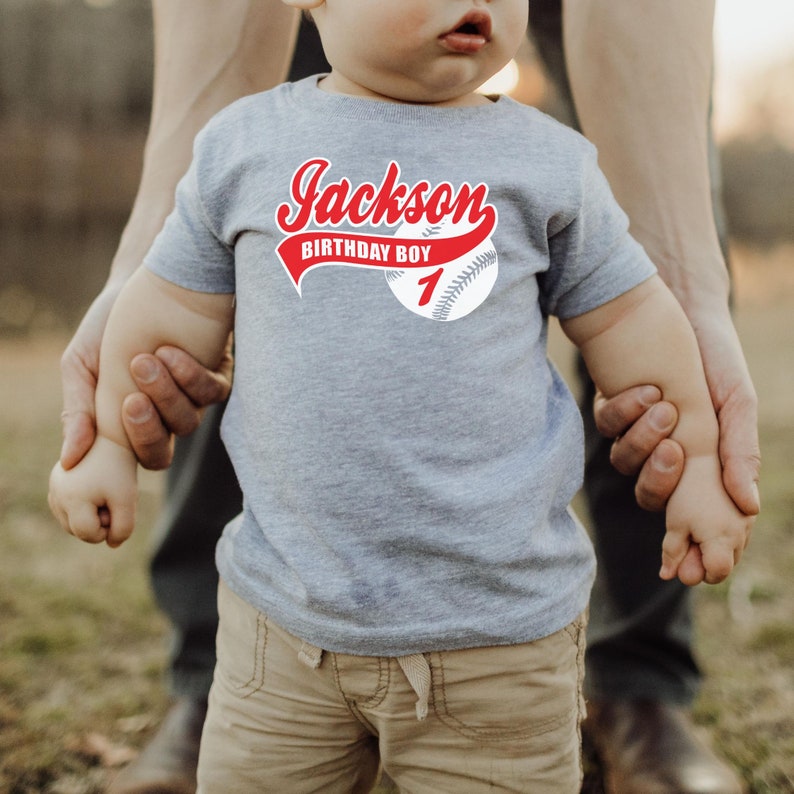 May include: A heather gray short-sleeve t-shirt featuring a graphic of a baseball with red text that reads Jackson, BIRTHDAY BOY, and the number 1. The design is centered on the chest of the shirt, which is worn by a young child.