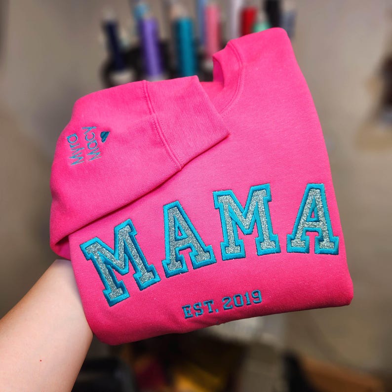 May include: A bright pink sweatshirt with the word "MAMA" embroidered in large, glittery, light blue letters. The text "EST. 2019" is embroidered below the word "MAMA". The sleeve cuff has the embroidered text "Myra Mccoy".