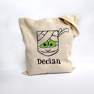 May include: A beige canvas tote bag featuring a mummy face design with green eyes and the name "Declan" in black. The mummy design is wrapped in bandages. The bag is made of a sturdy fabric.