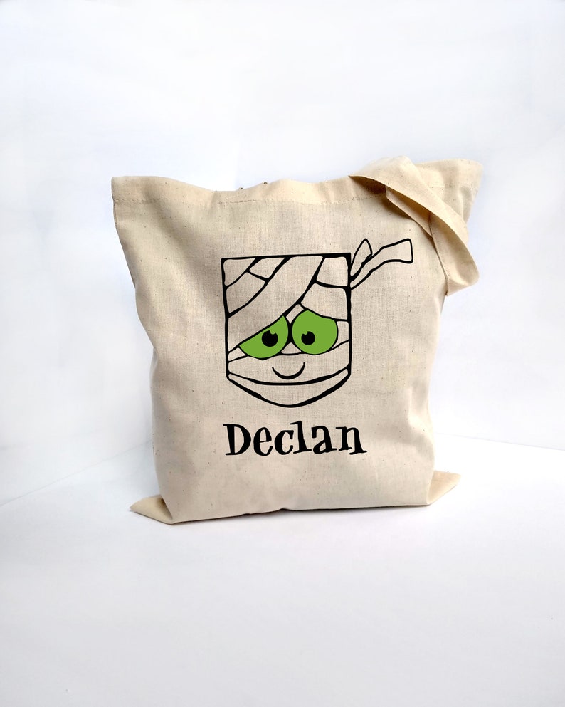 May include: A beige canvas tote bag featuring a mummy face design with green eyes and the name "Declan" in black. The mummy design is wrapped in bandages. The bag is made of a sturdy fabric.