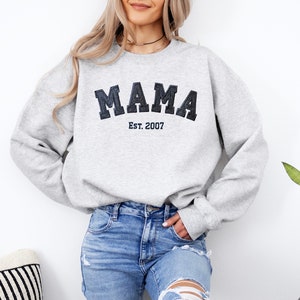 May include: A light gray sweatshirt with the word "MAMA" in dark blue, block letters, and "Est. 2007" below it. The sweatshirt has long sleeves and a crew neck. The model is wearing blue jeans.