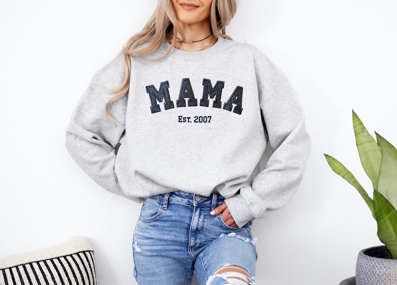 May include: A light gray sweatshirt with the word "MAMA" in dark blue, block letters, and "Est. 2007" below it. The sweatshirt has long sleeves and a crew neck. The model is wearing blue jeans.