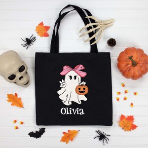 May include: Black tote bag with a Halloween ghost graphic holding a pumpkin and wearing a pink bow. The name "Olivia" is printed below the ghost. The image includes a pumpkin, skull, and fall-themed decorations.