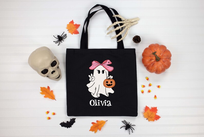 May include: Black tote bag with a Halloween ghost graphic holding a pumpkin and wearing a pink bow. The name "Olivia" is printed below the ghost. The image includes a pumpkin, skull, and fall-themed decorations.
