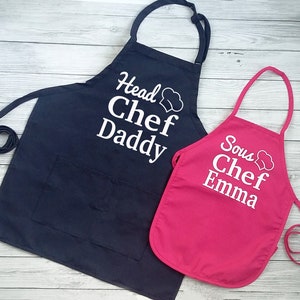 Peut inclure: Deux tabliers, un bleu marine et un rose vif, avec du texte blanc. Le tablier bleu marine dit "Head Chef Daddy" et le tablier rose dit "Sous Chef Emma". Les deux tabliers ont un motif de toque de chef.