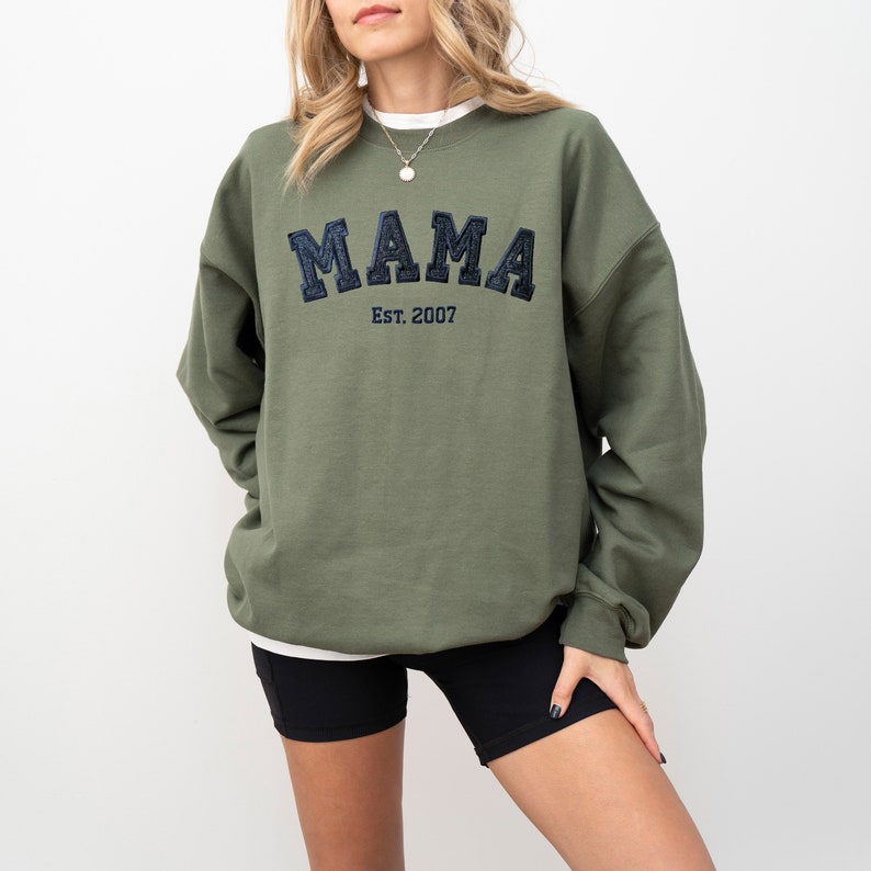 May include: Olive green sweatshirt with the word "MAMA" embroidered in navy blue, with "Est. 2007" underneath. The sweatshirt is a crew neck style.