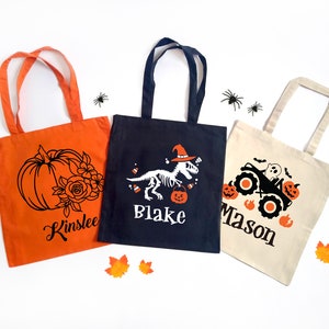 May include: Three Halloween-themed tote bags. An orange bag features a pumpkin and floral design with the name "Kinslee". A navy bag displays a dinosaur skeleton with the name "Blake". A beige bag has a monster truck design with the name "Mason".