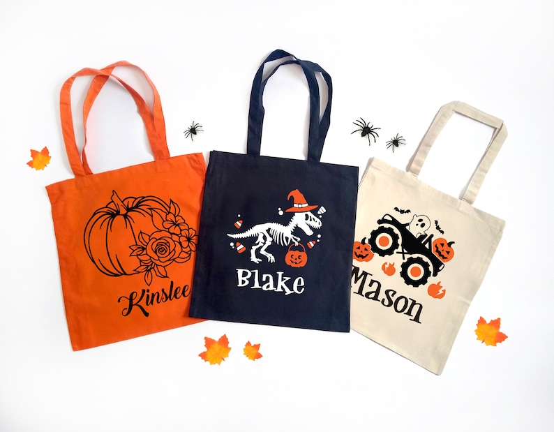 May include: Three Halloween-themed tote bags. An orange bag features a pumpkin and floral design with the name "Kinslee". A navy bag displays a dinosaur skeleton with the name "Blake". A beige bag has a monster truck design with the name "Mason".