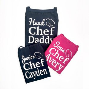 Peut inclure: Trois tabliers de cuisine, deux bleus marine et un magenta. Les tabliers bleus portent l'inscription blanche "Head Chef Daddy" et "Junior Chef Cayden". Le tablier magenta porte l'inscription blanche "Sous Chef Avery". Chaque tablier a un chapeau de chef.
