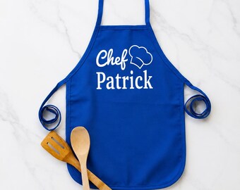 Könnte beinhalten: Eine königsblaue Schürze mit dem Text "Chef Patrick" und einer weißen Kochmützen-Grafik. Enthält einen Holzlöffel und einen Spatel. Die Schürze ist für das Kochen und die Zubereitung von Speisen bestimmt.