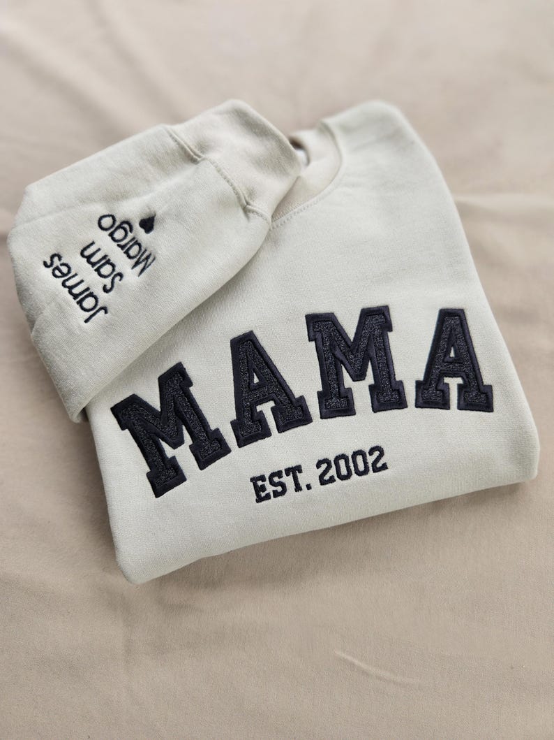 May include: Cream-colored sweatshirt with the word "MAMA" in navy blue varsity-style lettering. Below the word "MAMA" is the text "EST. 2002". The left sleeve has the names "James", "Sam", and "Margo" embroidered in navy blue.