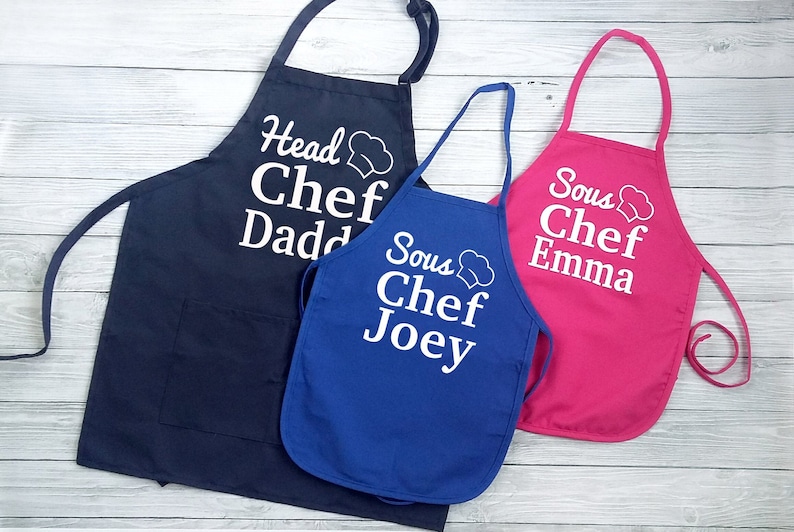 Peut inclure: Trois tabliers de cuisine : bleu marine, bleu roi et rose. Le tablier bleu marine porte l'inscription "Head Chef Dadd" avec un dessin de toque. Le bleu roi dit "Sous Chef Joey". Le rose dit "Sous Chef Emma".