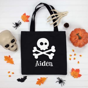 May include: Black tote bag with a white skull and crossbones design and the name "Aiden" printed below. The bag is surrounded by Halloween decorations, including a pumpkin, skull, and spiders.