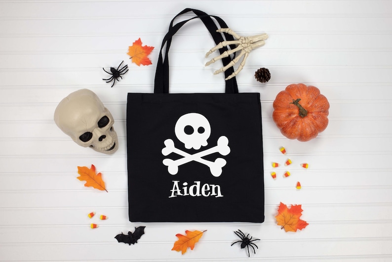 May include: Black tote bag with a white skull and crossbones design and the name "Aiden" printed below. The bag is surrounded by Halloween decorations, including a pumpkin, skull, and spiders.