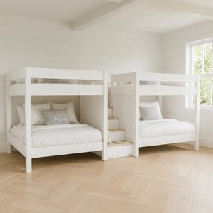 May include: A white wooden bunk bed set featuring two levels of sleeping space connected by a central staircase with built-in storage. Each bed is dressed with white bedding and a single gray patterned throw pillow, creating a clean, uniform look.