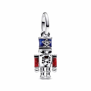 May include: A silver-colored charm in the shape of a nutcracker soldier. The charm features a blue and red color scheme with a star on the hat and red glitter accents on the arms. The charm has a loop at the top for attachment.