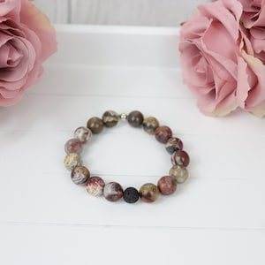 May include: A beaded bracelet featuring a mix of earth-toned jasper and lava rock beads. The bracelet has a silver-colored clasp and is displayed on a white surface with pink roses in the background. The beads are approximately 0.4 inches in diameter.