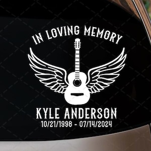 May include: A white vinyl decal applied to a car window. The design features a guitar with angel wings, the text "IN LOVING MEMORY," and the name "KYLE ANDERSON" with dates 10/21/1998 - 07/14/2024.