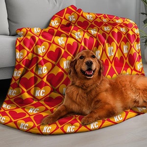 May include: A red and yellow fleece blanket with a repeating pattern of hearts and the letters "KC". A golden retriever dog is lying on the blanket.
