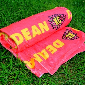 May include: A pink beach towel with the name "DEAN" written in yellow letters. The towel has a black and yellow leopard print arrow design with the letters "KC" in the center.